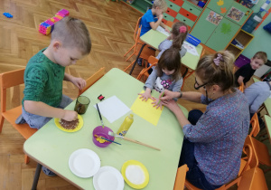 na zdjeciu chłopiec maluje papierowy talerzyk na zółto, obok niego siedzi dziewczynka , której pani odrysowuje dłonie nanżółtym kartonie, na stoliku leżą pędzelki, talerzyki i żółta farba- materiały potrzebne do wykonania pracy.